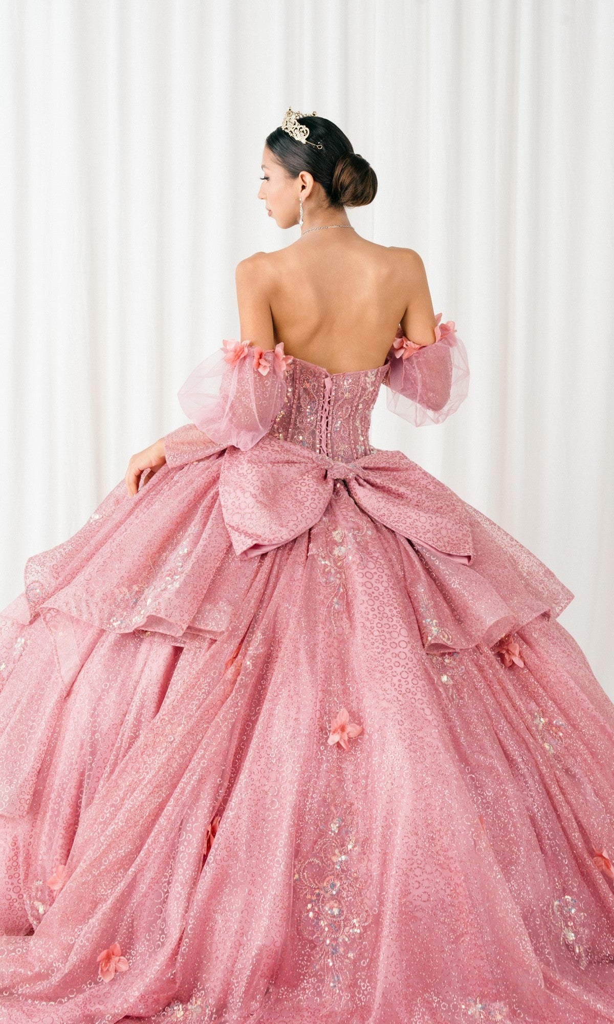 A woman faces away in the Quinceanera Dress 8385J By Cinderella Couture, a dusty rose gown with off-shoulder, corset bodice, floral details, and a large bow. Her bun with tiara completes the look against a white backdrop.